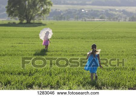 Stock Photograph - A young girl running to catch up with her twin sister in sunny field. Fotosearch