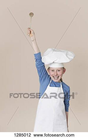 Stock Image - An excited girl wearing a chef's hat and apron holding a wooden spoon up. Fotosearch