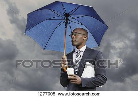 Businessman holding umbrella, disposable coffee cup and newspaper under a moody sky View Large Photo Image Stock Photo - Businessman holding umbrella, disposable coffee cup and newspaper under a moody sky. Fotosearch