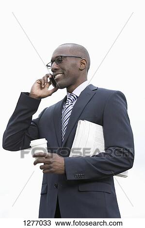Stock Image - Businessman with newspaper underarm holding disposable coffee cup and talking on phone. Fotosearch