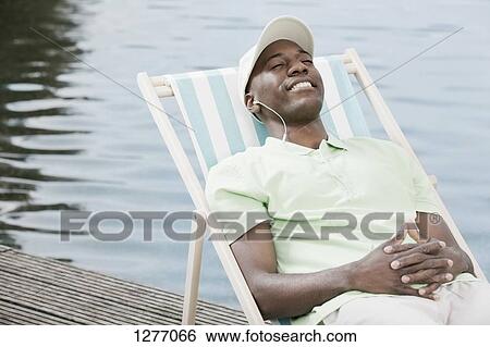 Stock Photograph - Happy man relaxing on deck chair by lake wearing headphones. Fotosearch