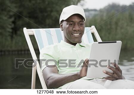 Stock Photo - Man on deck chair using digital tablet by lake. Fotosearch