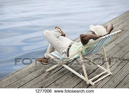 Man relaxing by lake on deck chair View Large Photo Image Stock Photograph - Man relaxing by lake on deck chair. Fotosearch