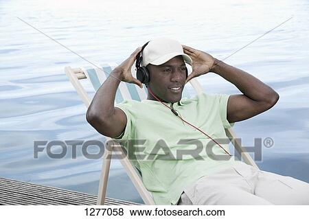 Man relaxing on deck chair by lake wearing headphones View Large Photo Image Stock Photo - Man relaxing on deck chair by lake wearing headphones. Fotosearch