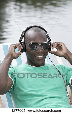 Stock Photography - Man relaxing on deck chair by lake wearing headphones. Fotosearch