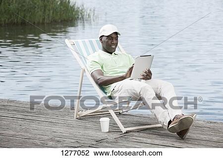 Man relaxing on deck chair using digital tablet by lake View Large Photo Image Stock Image - Man relaxing on deck chair using digital tablet by lake. Fotosearch