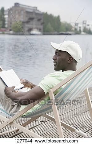 Picture - Man relaxing with book on deck chair by lake. Fotosearch