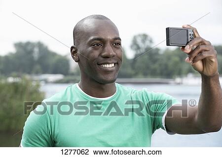 Stock Image - Smiling man taking photograph of himself by lake. Fotosearch