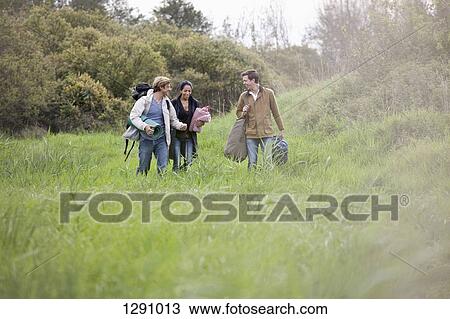 Stock Image - Men and woman carrying camping accessories and walking through filed. Fotosearch
