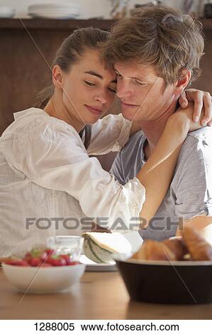Stock Photography - Young couple sitting at dining table, embracing. Fotosearch