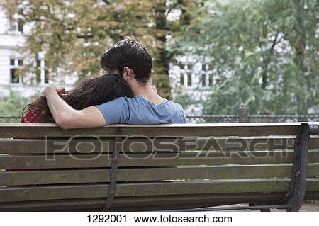 Stock Image - Young couple sitting on park bench, rear view. Fotosearch
