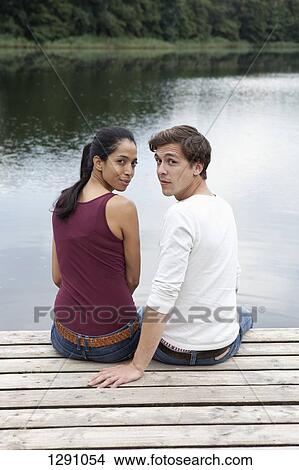 Picture - Young couple sitting on pier, looking over shoulders. Fotosearch