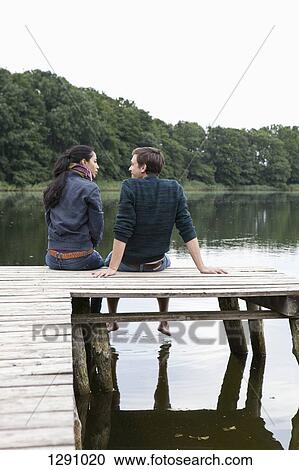 Stock Image - Young couple sitting on wooden pier. Fotosearch