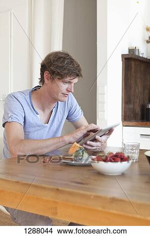 Young man sitting at dining table using digital tablet View Large Photo Image Picture - Young man sitting at dining table using digital tablet. Fotosearch