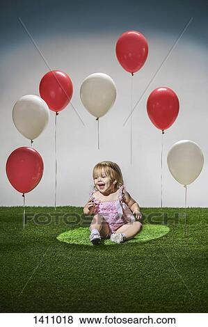 Stock Photo - Cheerful girl sitting on grass with red and white balloons in mid-air. Fotosearch