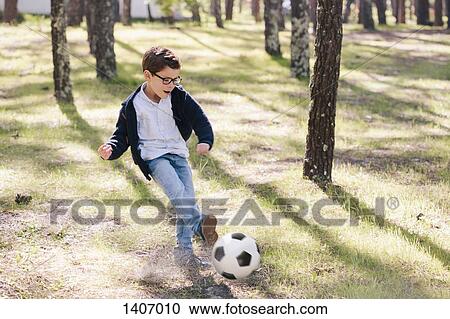 Stock Image - Full length of happy boy playing soccer in forest. Fotosearch