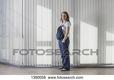 Stock Photo - Full length portrait of pregnant woman in overalls standing against curtain. Fotosearch