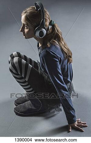 Picture - Full length side view of girl crouching while listening music through headphones over gray backgroun. Fotosearch