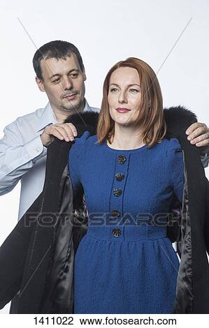 Stock Image - Man assisting woman in wearing winter coat against white background. Fotosearch