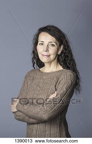Stock Image - Portrait of confident mature woman standing arms crossed over gray background. Fotosearch