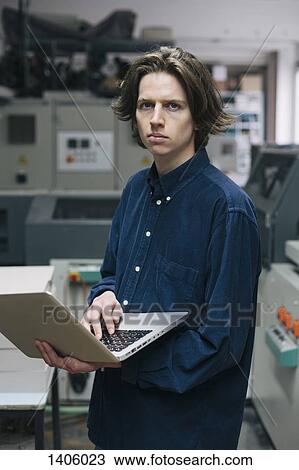 Stock Image - Portrait of confident young male technician using laptop in factory. Fotosearch
