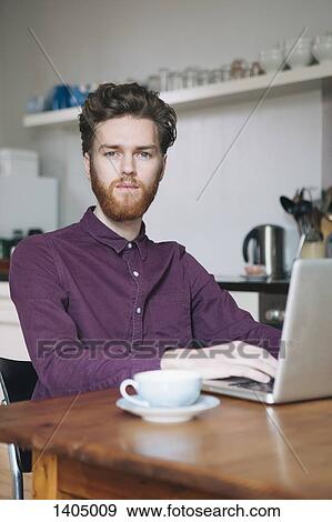 Stock Photo - Portrait of confident young man using laptop at table in kitchen. Fotosearch