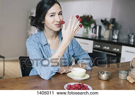 Stock Image - Portrait of happy young woman holding raspberries on fingers at home. Fotosearch