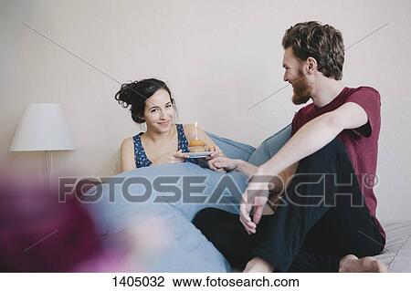 Stock Image - Portrait of happy young woman holding birthday cupcake with candle while sitting beside man on bed. Fotosearch