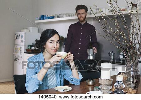 Portrait of young woman having coffee at kitchen table with man in background View Large Photo Image Stock Image - Portrait of young woman having coffee at kitchen table with man in background. Fotosearch