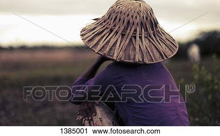 Stock Image - "Rear view of woman in bamboo hat sitting on field, Bago region, Myanmar". Fotosearch