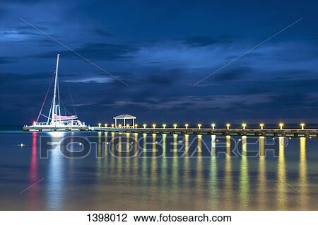 Stock Image - Sailboat moored at illuminated pier on lake against sky at night. Fotosearch
