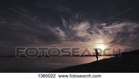 Stock Image - "Silhouette man standing at lake shore during sunset, Aydar lake, Uzbekistan". Fotosearch