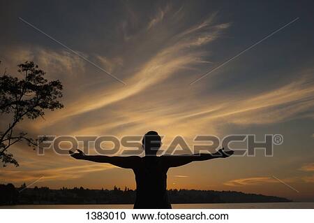 Stock Image - Silhouette young woman standing with arms outstretched on lakeshore during sunset. Fotosearch
