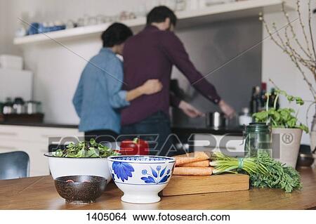 Vegetables on kitchen table with couple cooking in background View Large Photo Image Picture - Vegetables on kitchen table with couple cooking in background. Fotosearch