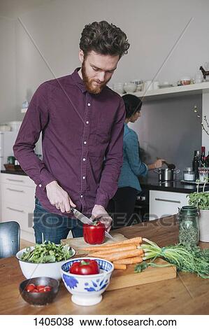 Stock Photo - Young man chopping red bell pepper at kitchen table with woman in background. Fotosearch