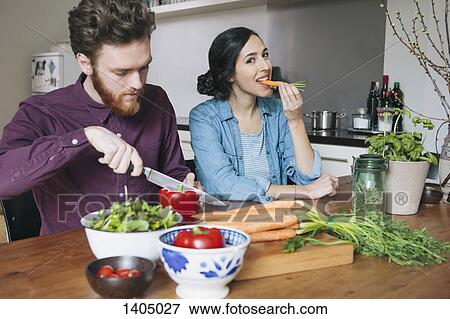Young man chopping red bell pepper beside woman eating carrot at kitchen table View Large Photo Image Stock Photo - Young man chopping red bell pepper beside woman eating carrot at kitchen table. Fotosearch