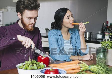Stock Photo - Young man chopping red bell pepper beside woman eating carrot at kitchen table. Fotosearch