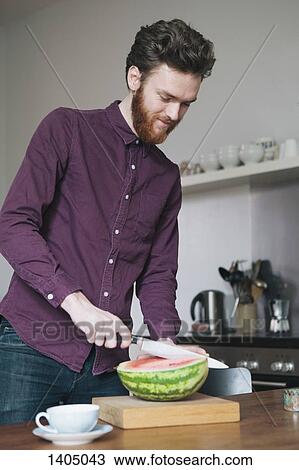 Stock Image - Young man cutting watermelon on cutting board in kitchen. Fotosearch