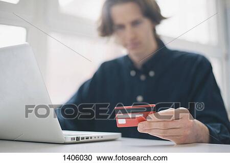 Stock Image - Young man holding credit card while using laptop at a desk. Fotosearch