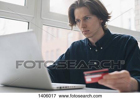 Young man holding credit card while using laptop at table View Large Photo Image Stock Photo - Young man holding credit card while using laptop at table. Fotosearch