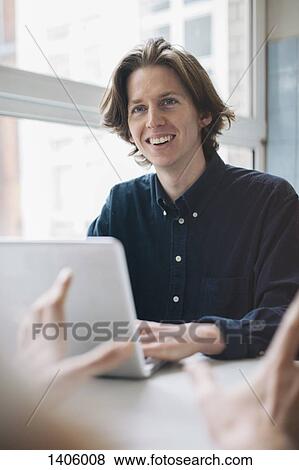 Young man smiling at colleague while using laptop at table View Large Photo Image Stock Photo - Young man smiling at colleague while using laptop at table. Fotosearch
