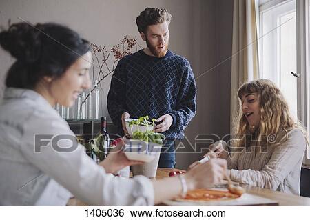 Stock Photograph - Young man with female friends making pizza at table. Fotosearch