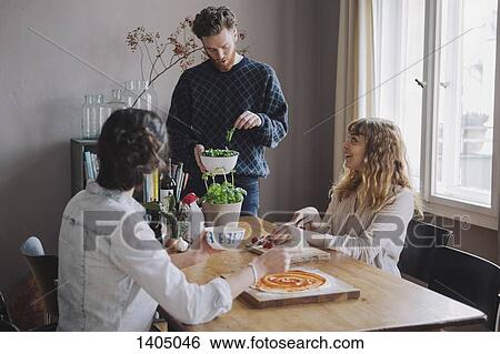 Stock Photograph - Young man with female friends making pizza at table. Fotosearch