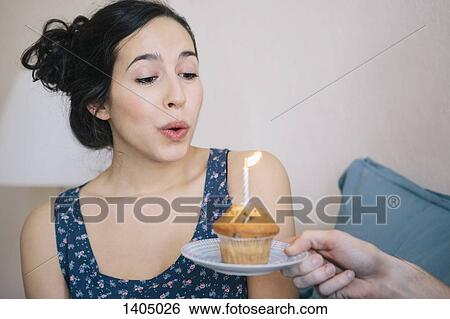 Stock Photograph - Young woman blowing out candle on cupcake held by man at home. Fotosearch