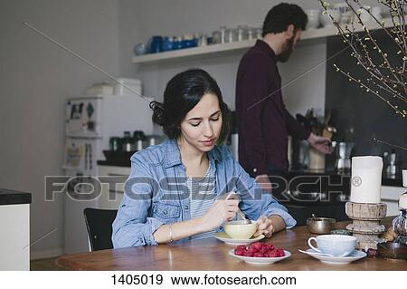 Stock Photo - Young woman stirring coffee at kitchen table with man in background. Fotosearch