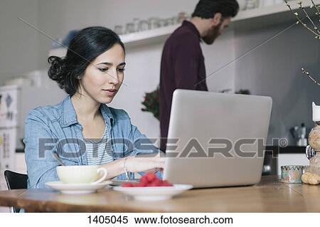 Stock Photography - Young woman using laptop with man in background at kitchen. Fotosearch
