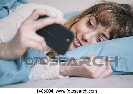 Young woman using mobile phone while relaxing in bed View Large Photo Image Picture - Young woman using mobile phone while relaxing in bed. Fotosearch
