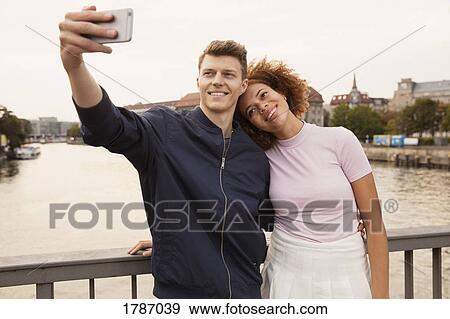 Stock Photo - Affectionate young couple with smart phone taking selfie on urban bridge. Fotosearch