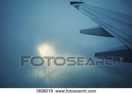 Stock Photo - Airplane wing flying through clouds over water. Fotosearch