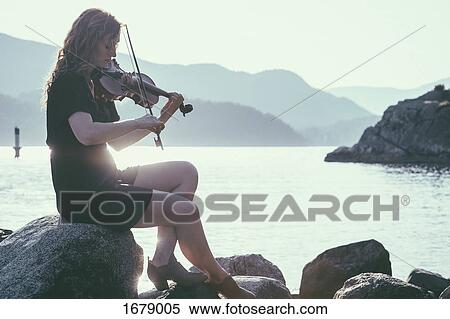 Stock Photography - Backlit woman playing violin while sitting on rock at lakeshore against mountains. Fotosearch
